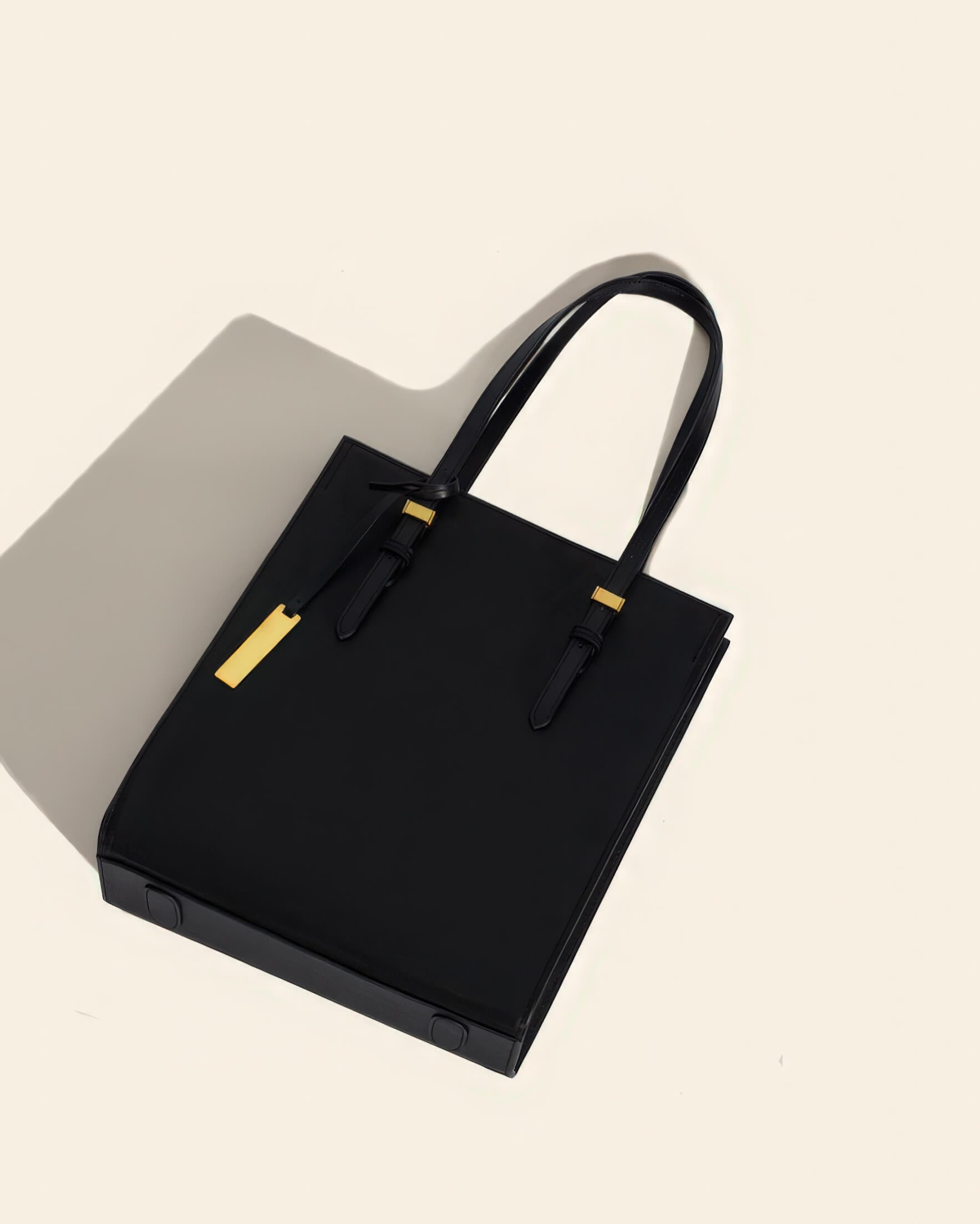 Flat lay of a minimalist black leather tote bag with adjustable straps and gold accent hardware.
