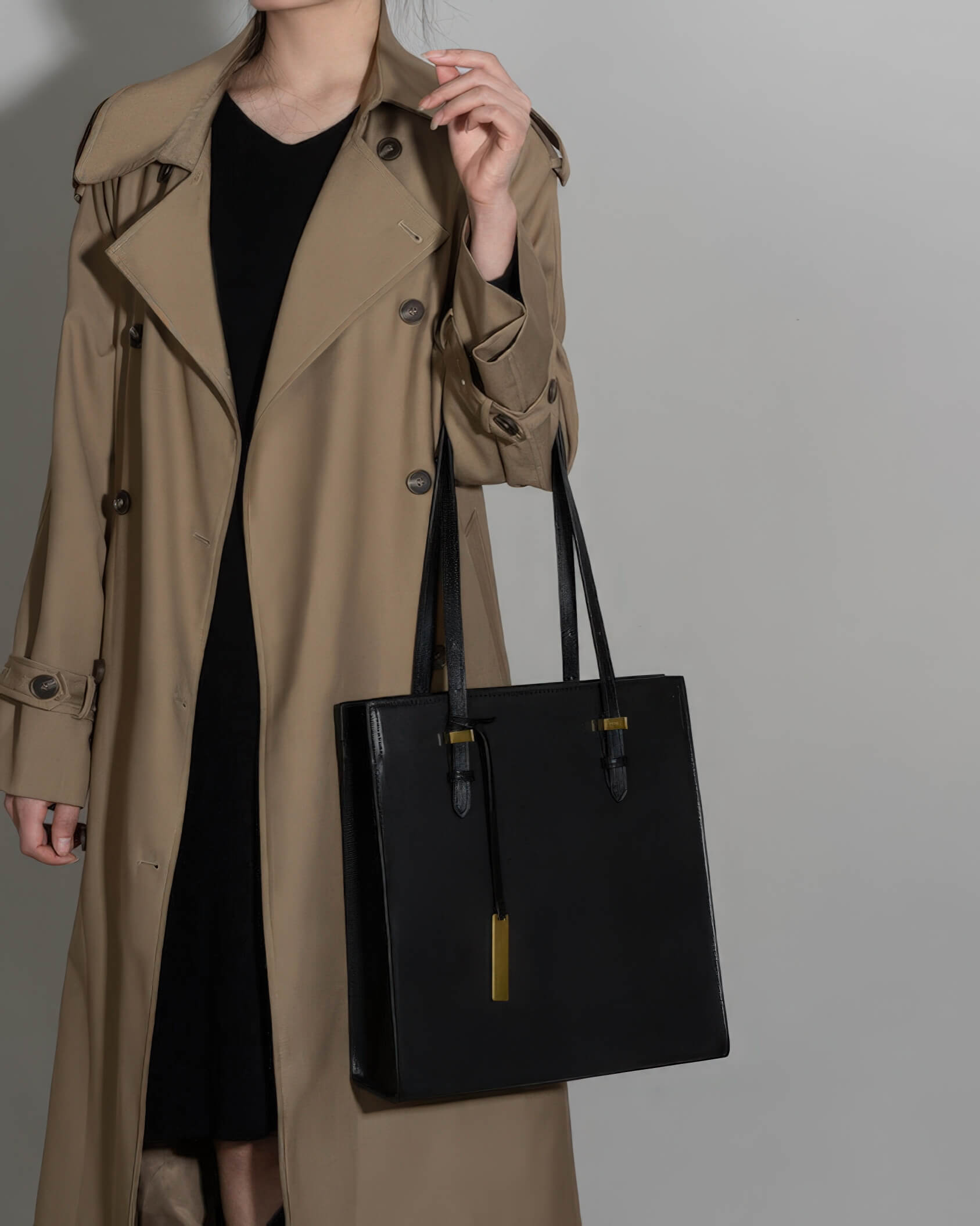 Woman in a tan trench coat holding a structured black leather tote bag by her side.