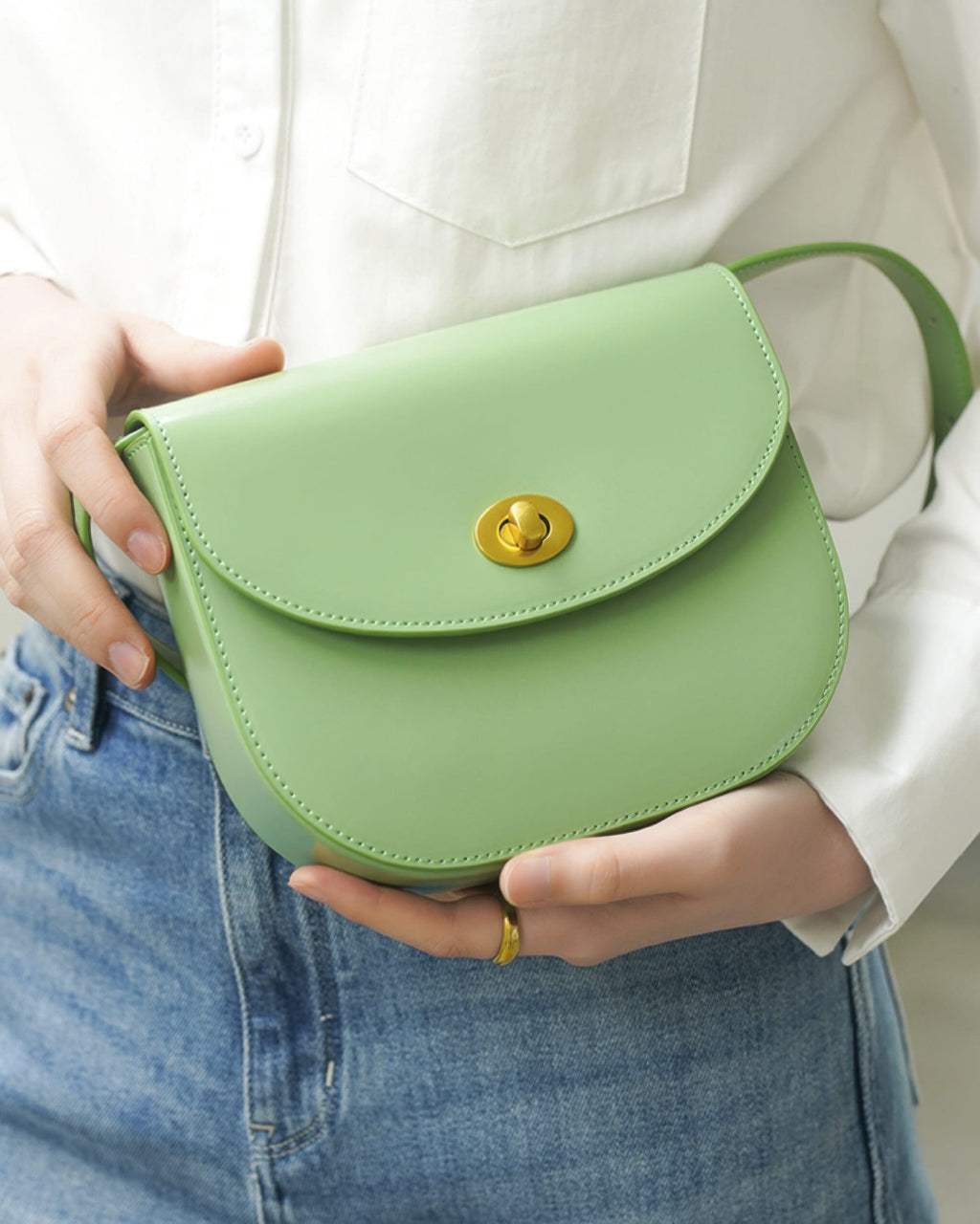 Close-up of a model holding a sage green classic leather flap saddle crossbody bag while wearing a white shirt and blue jeans.