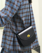 Model wearing a blue plaid jacket carrying a black leather flap saddle crossbody bag with a gold turn-lock closure.