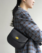 Model wearing a blue plaid jacket holding a black leather saddle bag over her shoulder.