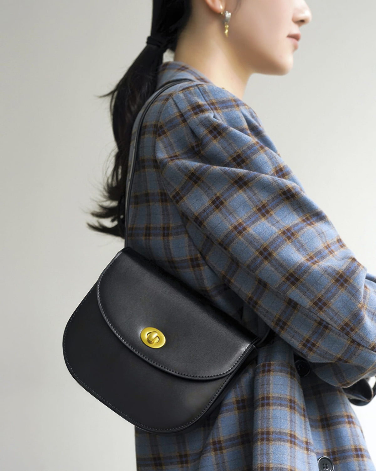 Model wearing a blue plaid jacket holding a black leather saddle bag over her shoulder.