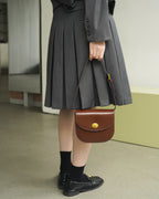 Model wearing a grey pleated skirt holding a dark brown leather flap saddle handbag.