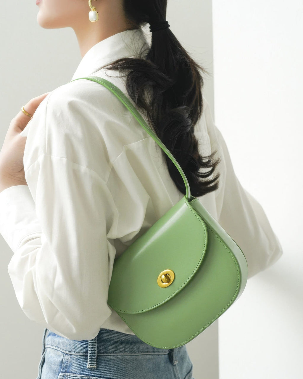 Back view of a model in a white button-up shirt wearing a sage green leather flap saddle bag over her shoulder.