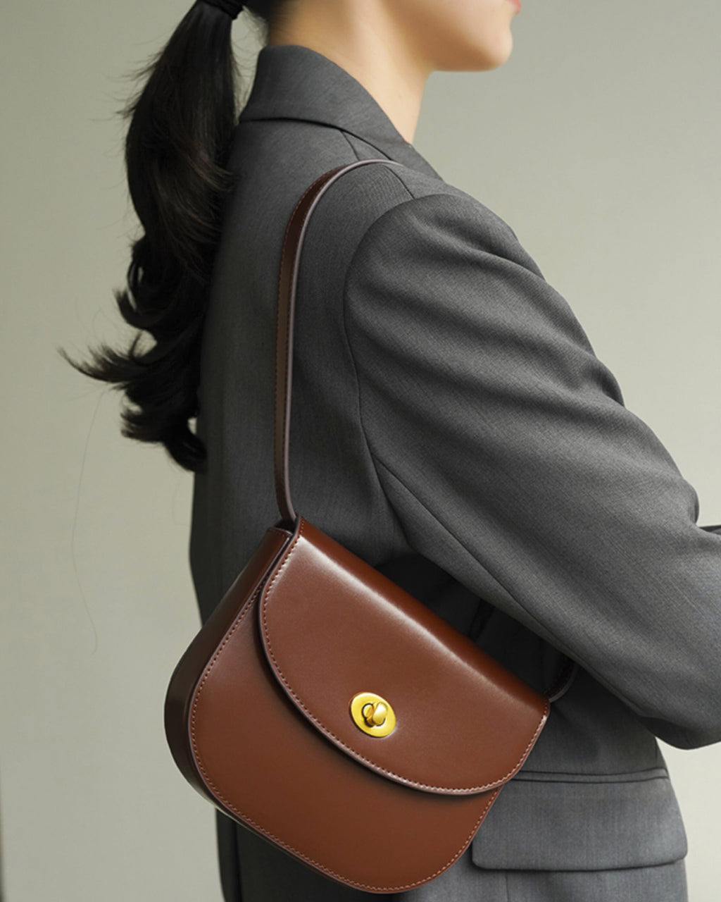Side profile of a brown classic leather flap saddle crossbody bag worn over the shoulder with a grey blazer.