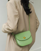 Back view of a sage green classic leather flap saddle crossbody bag worn by a model in a beige blazer.