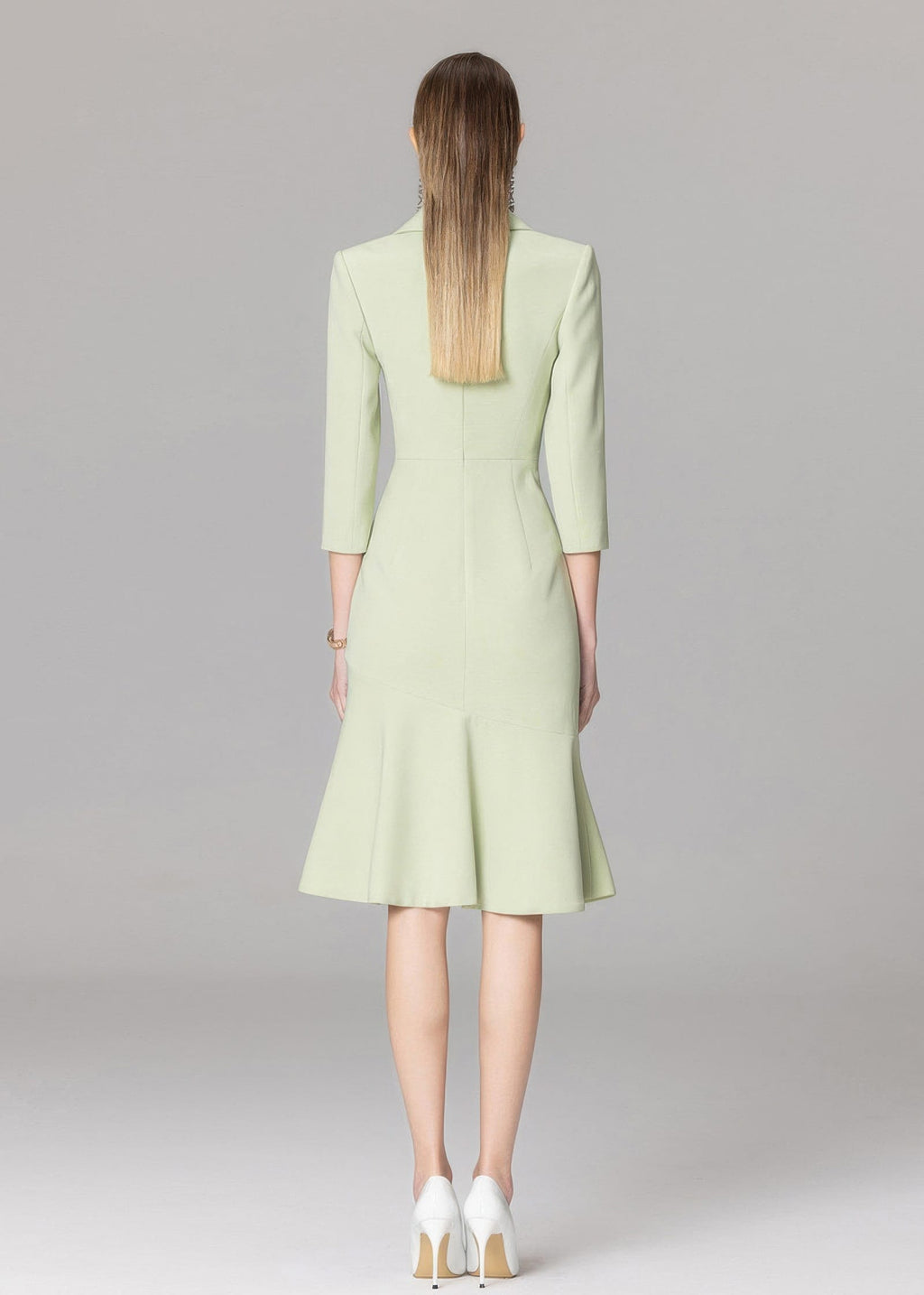 The back view of a model wearing a light green blazer dress, highlighting the fitted waist, mermaid-style skirt, and white high-heeled pumps.