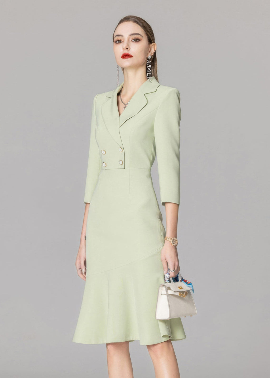 A three-quarter view of a model wearing a light green, double-breasted blazer dress with a mermaid hem, holding a small white handbag.