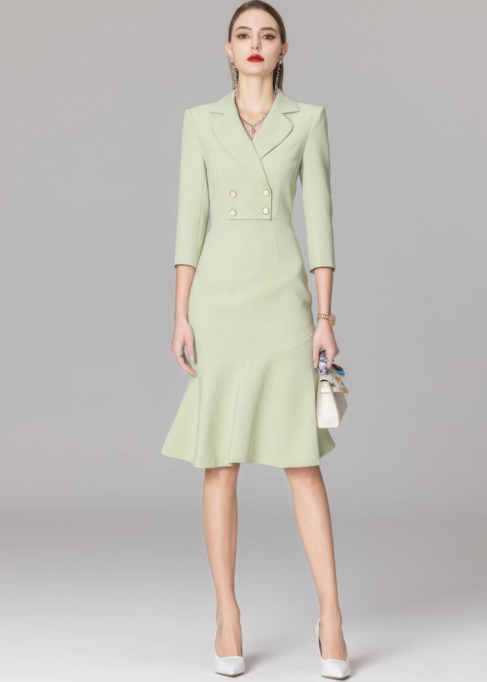 A full-length shot of a model in a light green blazer dress, holding a white top-handle bag with a silk scarf tied to it.