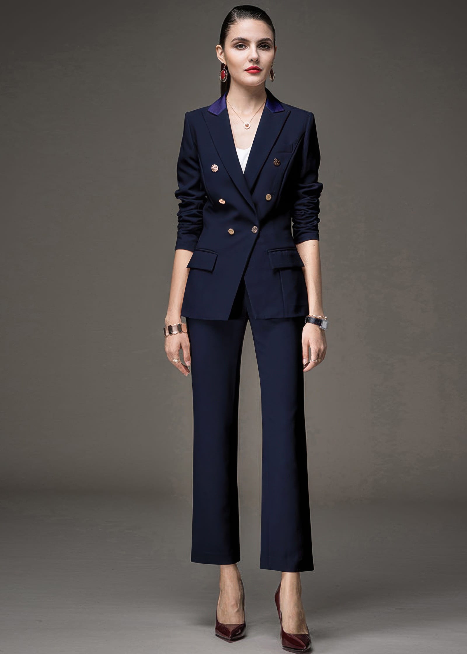 Full-length front view of a woman wearing a navy blue double-breasted pant suit with gold buttons and dark red heels.