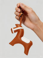 Hand holding an equestrian horse bag charm crafted from orange French goatskin leather with white saddle details.
