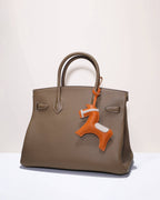 Full view of a stylish taupe tote bag decorated with an orange equestrian goatskin leather horse charm.