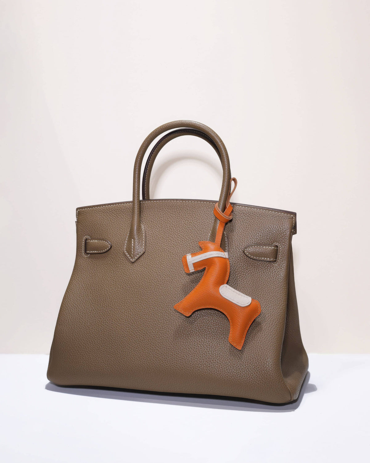Full view of a stylish taupe tote bag decorated with an orange equestrian goatskin leather horse charm.