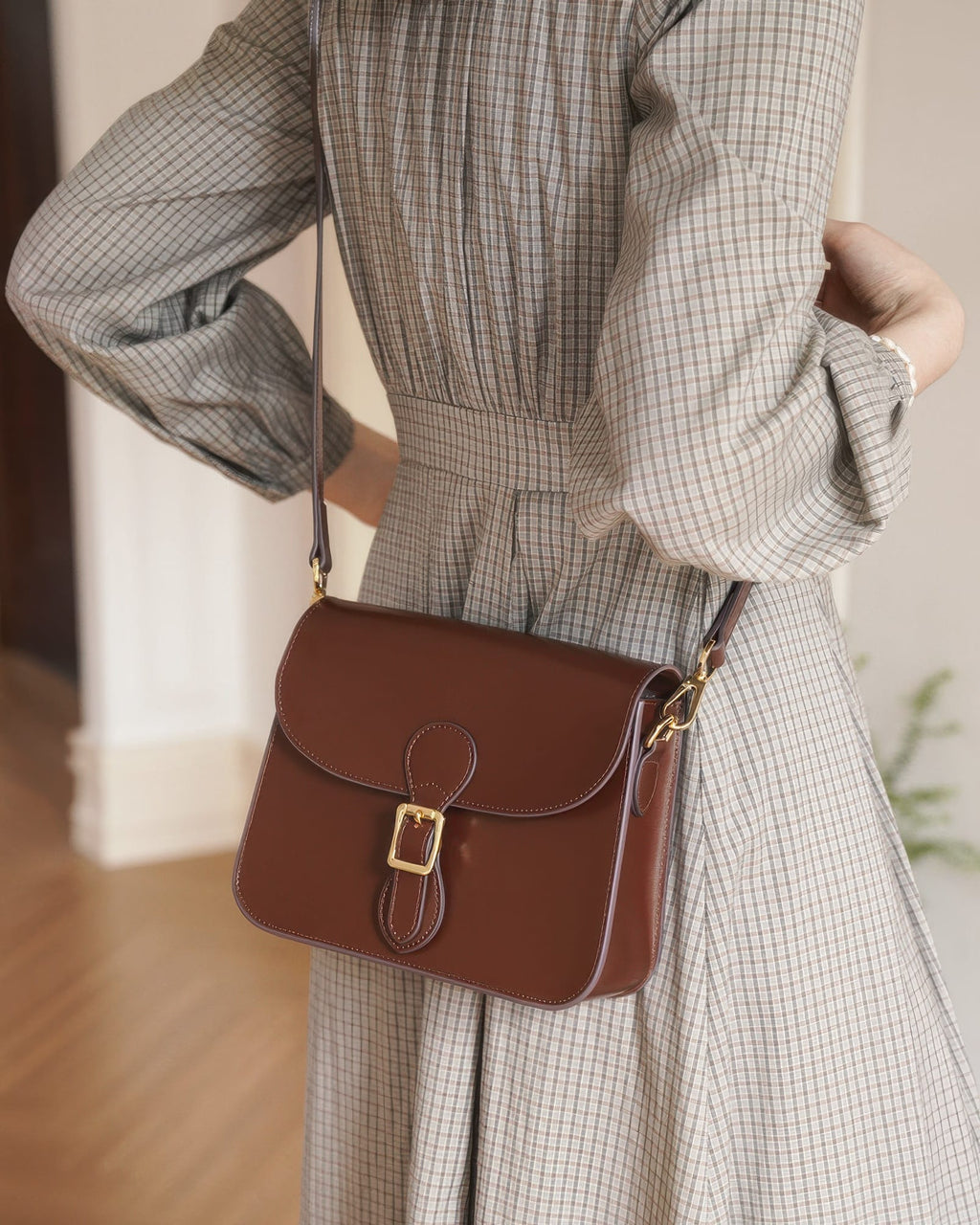Model wearing a plaid grey dress carrying a dark brown leather flap crossbody bag with a gold buckle detail.