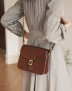 Model wearing a plaid grey dress carrying a dark brown leather flap crossbody bag with a gold buckle detail.