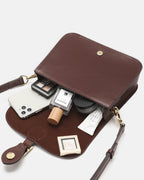 Open top-down interior view of a brown leather flap crossbody bag filled with daily essentials including a phone, perfume, and cosmetics.
