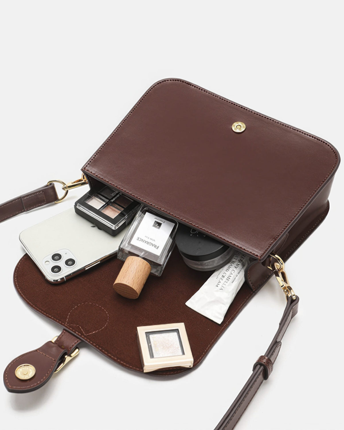 Open top-down interior view of a brown leather flap crossbody bag filled with daily essentials including a phone, perfume, and cosmetics.

