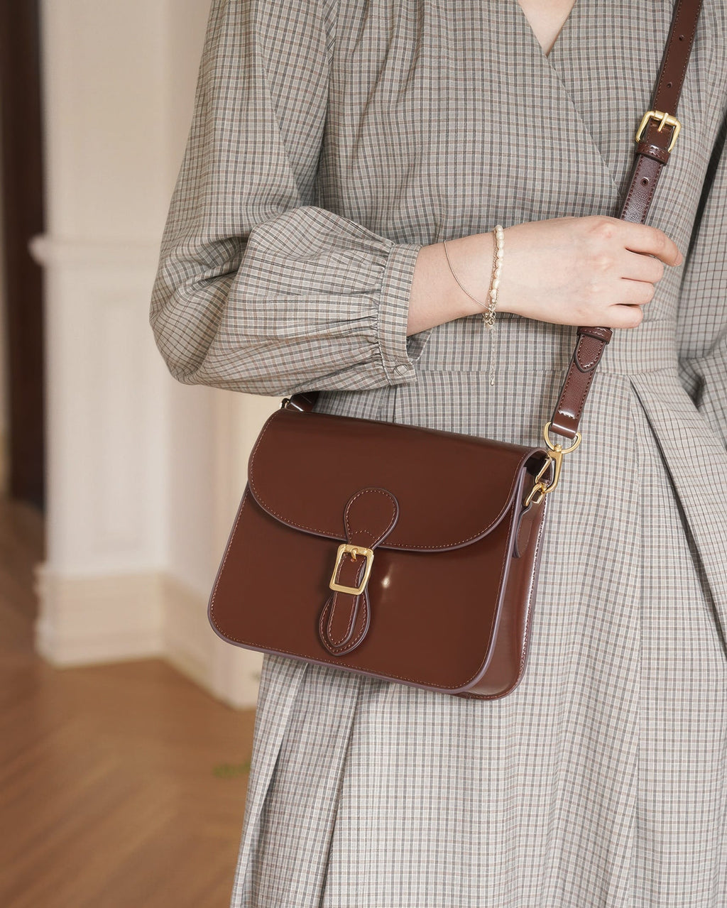 Front view of a model in a plaid dress wearing a dark brown leather flap crossbody bag with gold buckle detail.