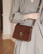 Front view of a model in a plaid dress wearing a dark brown leather flap crossbody bag with gold buckle detail.