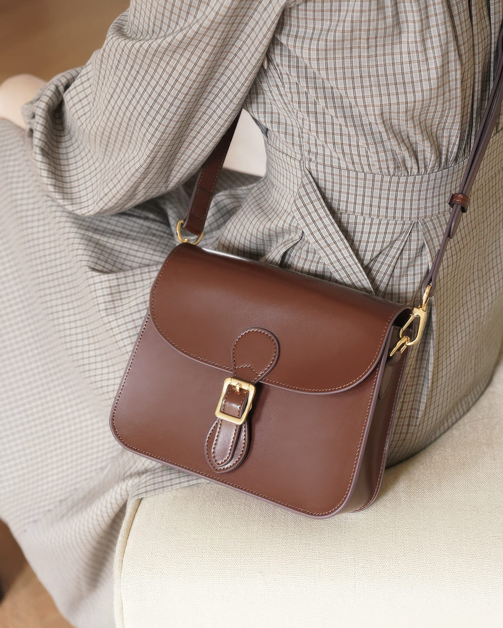 High-angle lifestyle view of a dark brown leather flap crossbody bag resting on a beige chair beside a model in a plaid dress.