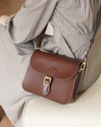 High-angle lifestyle view of a dark brown leather flap crossbody bag resting on a beige chair beside a model in a plaid dress.