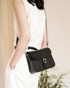Side view of a model in a white outfit wearing a black leather buckle flap crossbody bag.
