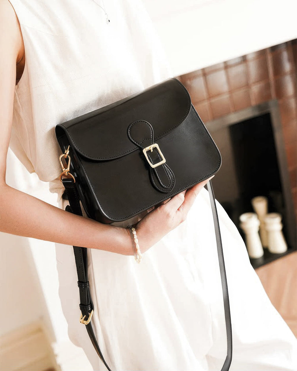 Front three-quarter angle of a model holding a black leather flap crossbody bag with gold buckle hardware.