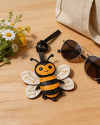 Honeybee leather bag charm displayed on a wooden table with sunglasses and white daisies.