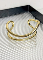 A single gold-tone fluid wave statement bangle showcasing its open-cuff design and smooth, curved lines.