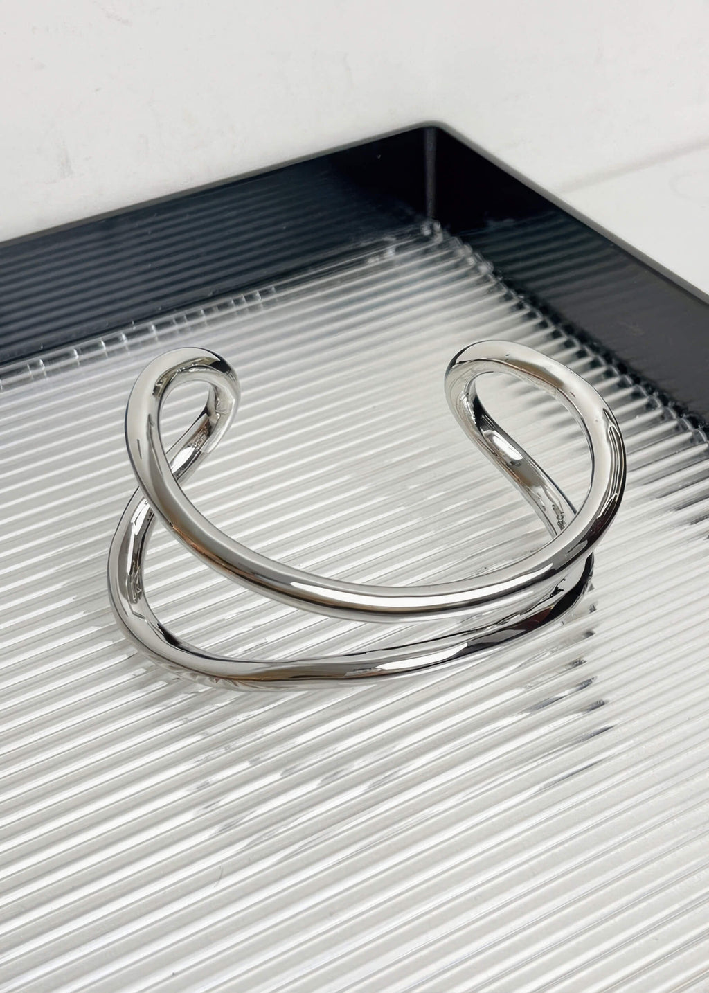 A single silver-tone sculptural metal bangle with a polished finish, displayed on a modern ribbed surface.