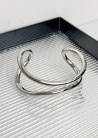 A single silver-tone sculptural metal bangle with a polished finish, displayed on a modern ribbed surface.