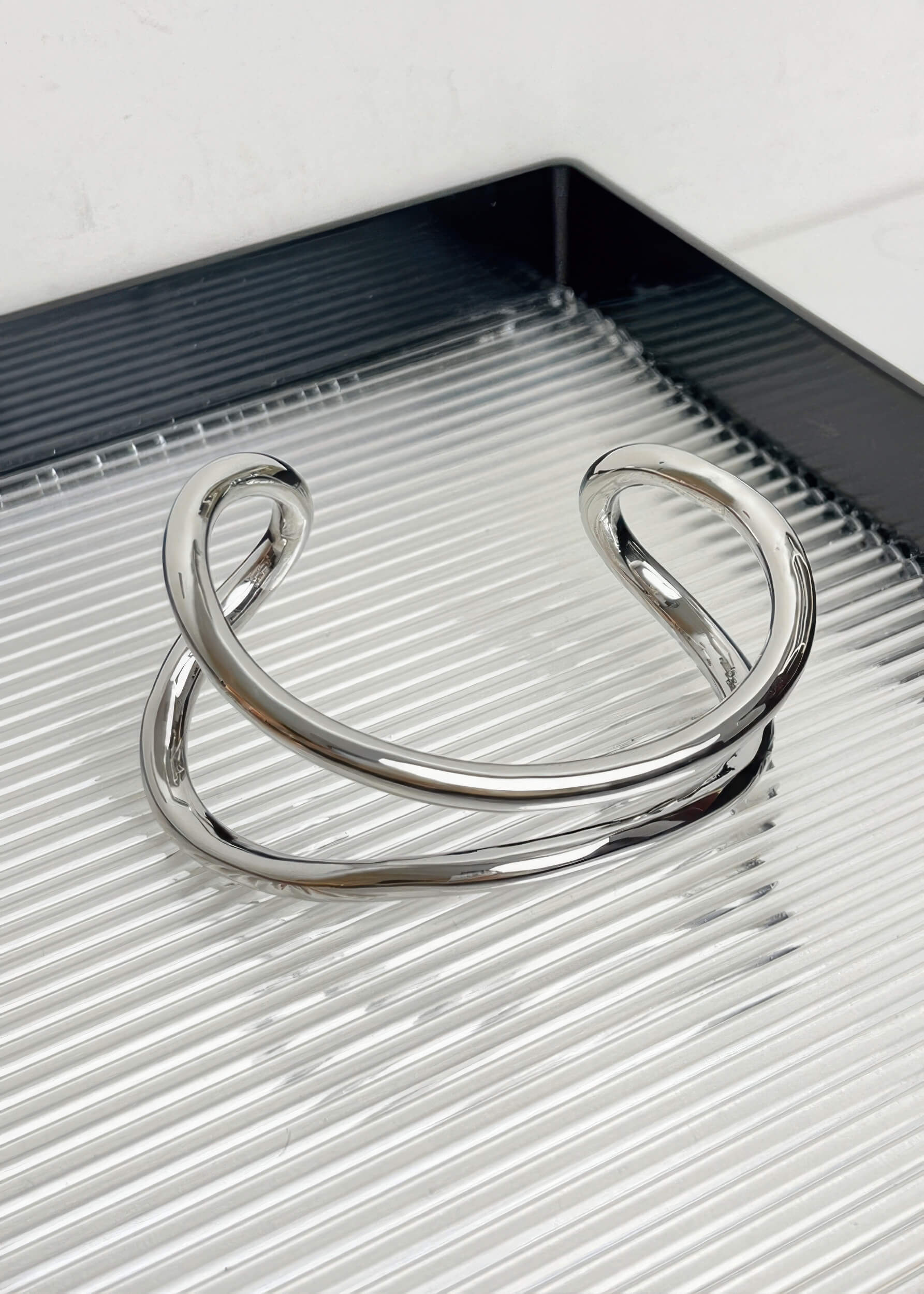 A single silver-tone sculptural metal bangle with a polished finish, displayed on a modern ribbed surface.