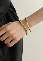 Close-up of a person wearing a gold minimalist fluid wave statement bangle on their wrist over a black dress.