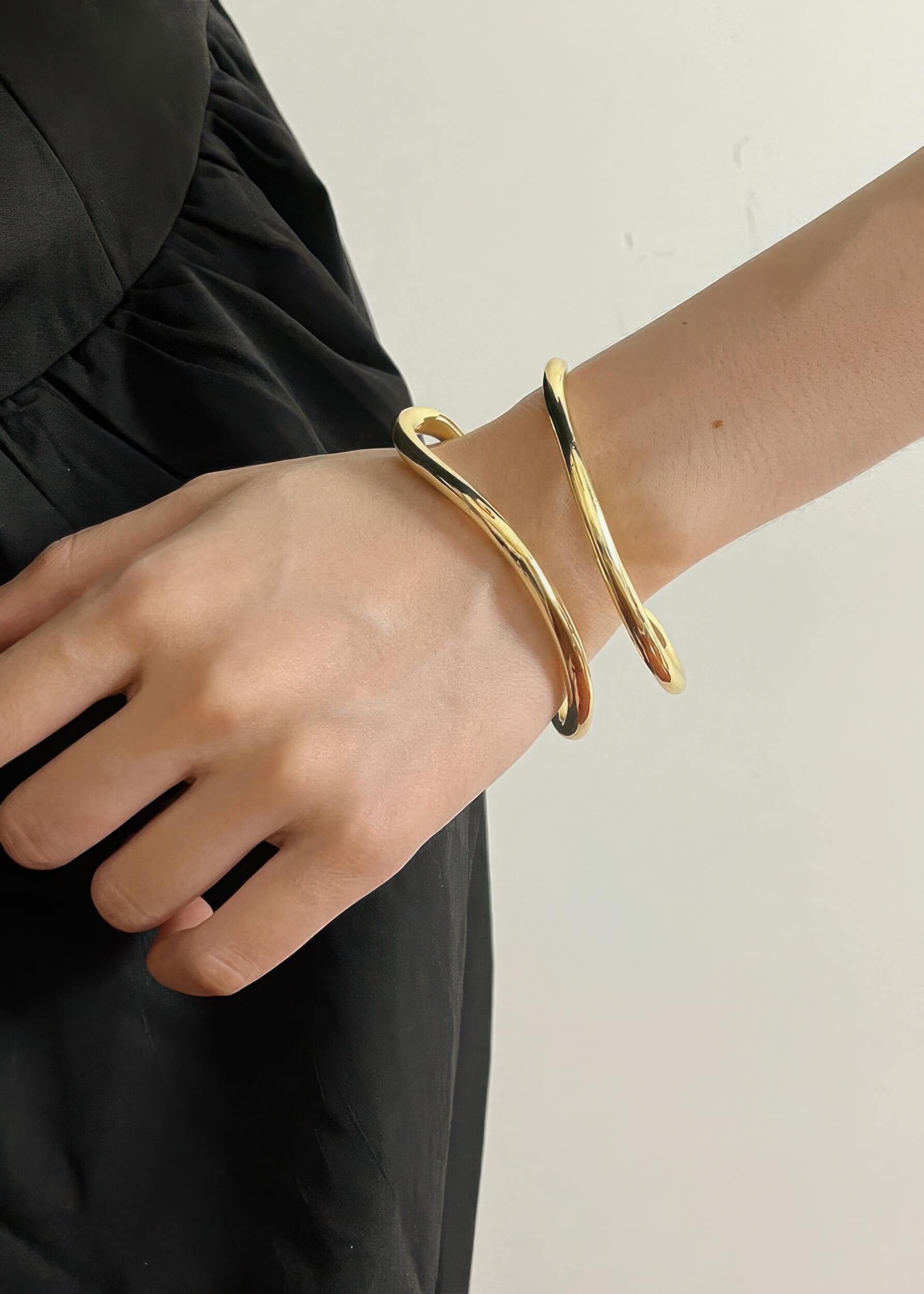 Close-up of a person wearing a gold minimalist fluid wave statement bangle on their wrist over a black dress.