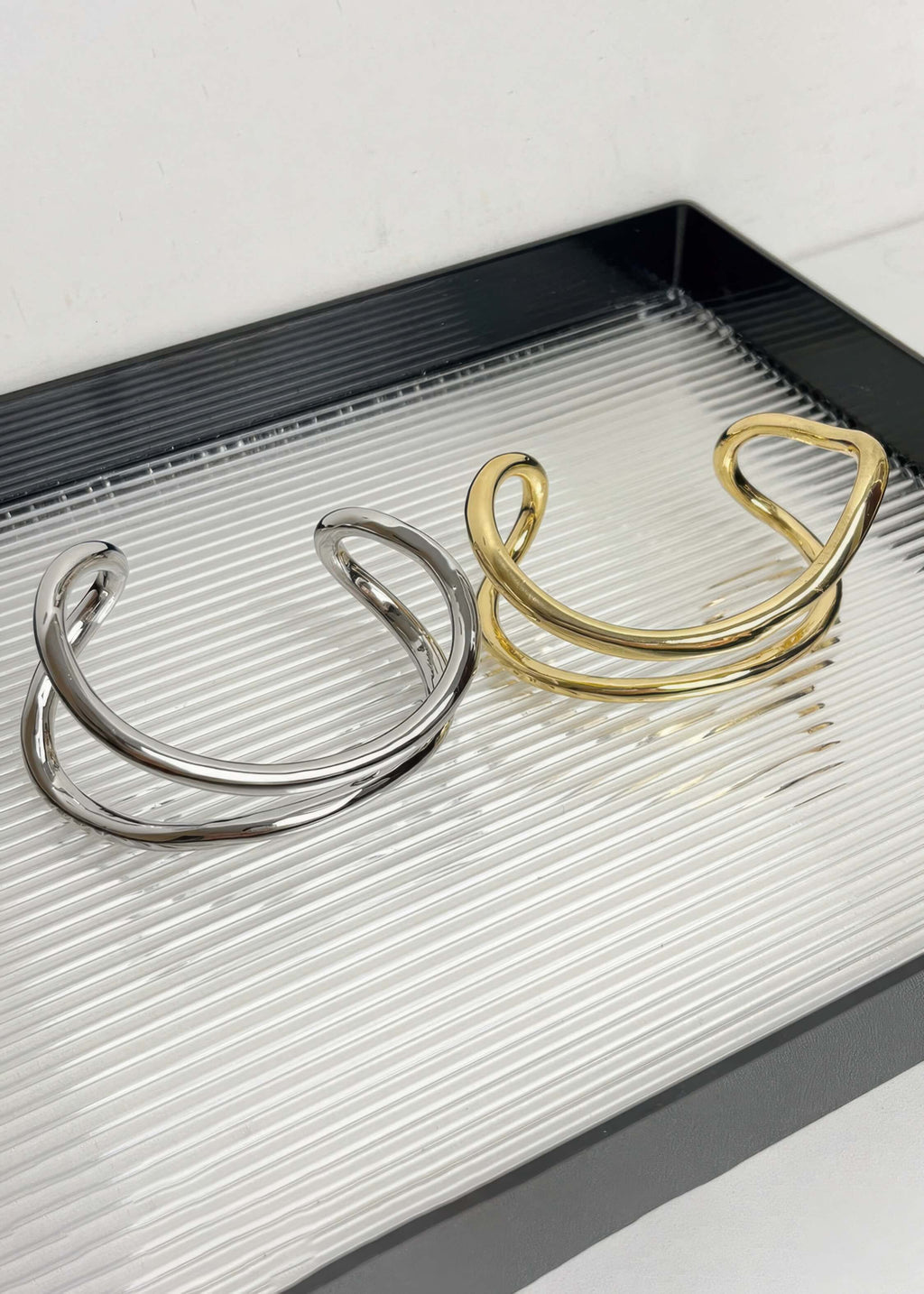 Two minimalist fluid wave bangles, one in polished gold and one in silver, displayed on a textured glass tray.