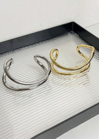 Two minimalist fluid wave bangles, one in polished gold and one in silver, displayed on a textured glass tray.