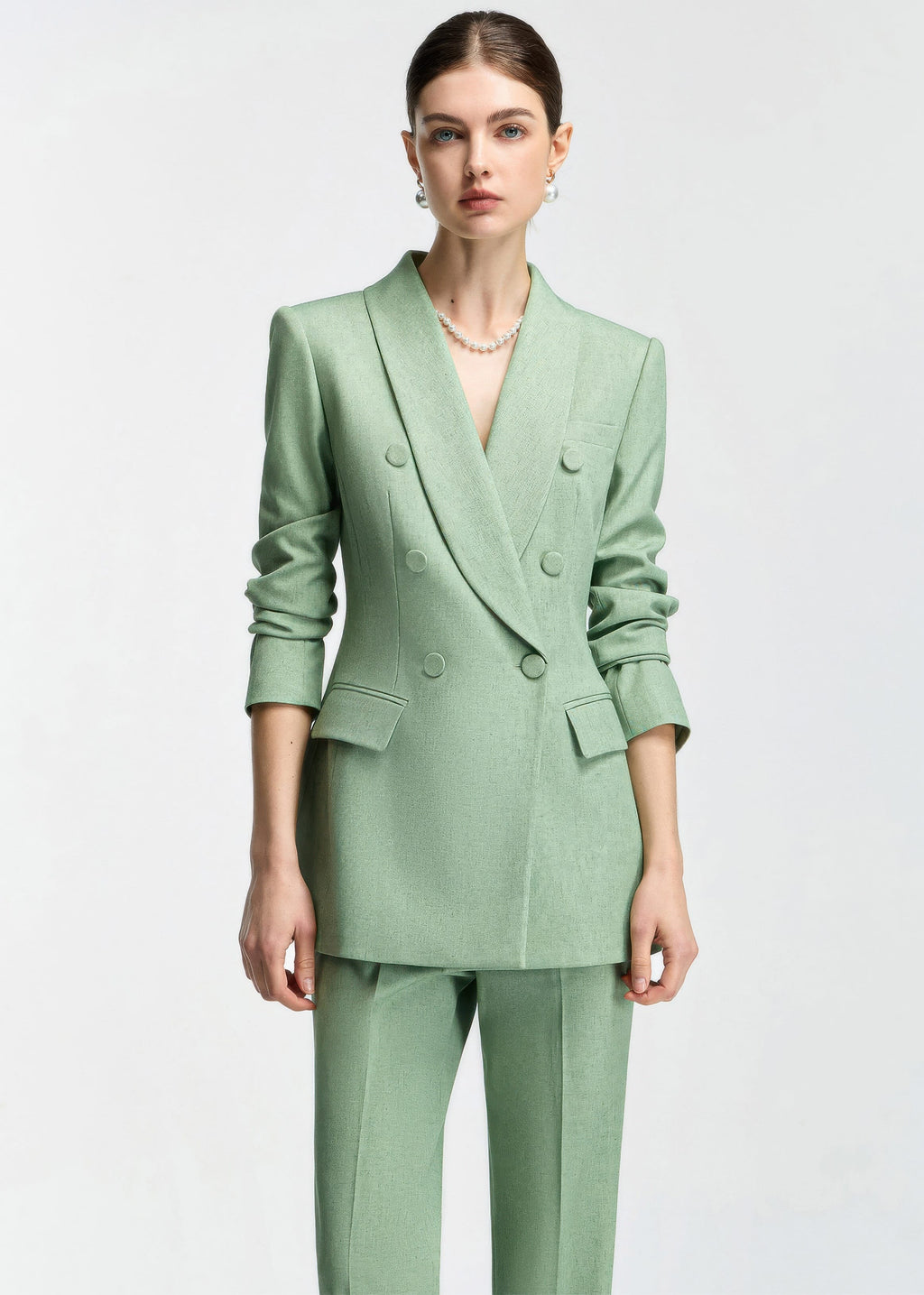 A front-facing portrait of a model in a mint green slim-fit pantsuit with sleeves pushed up, featuring double-breasted buttons and a pearl necklace.