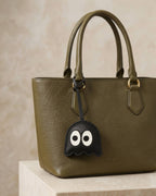 Black ghost leather bag charm attached to an olive green leather tote bag handle.