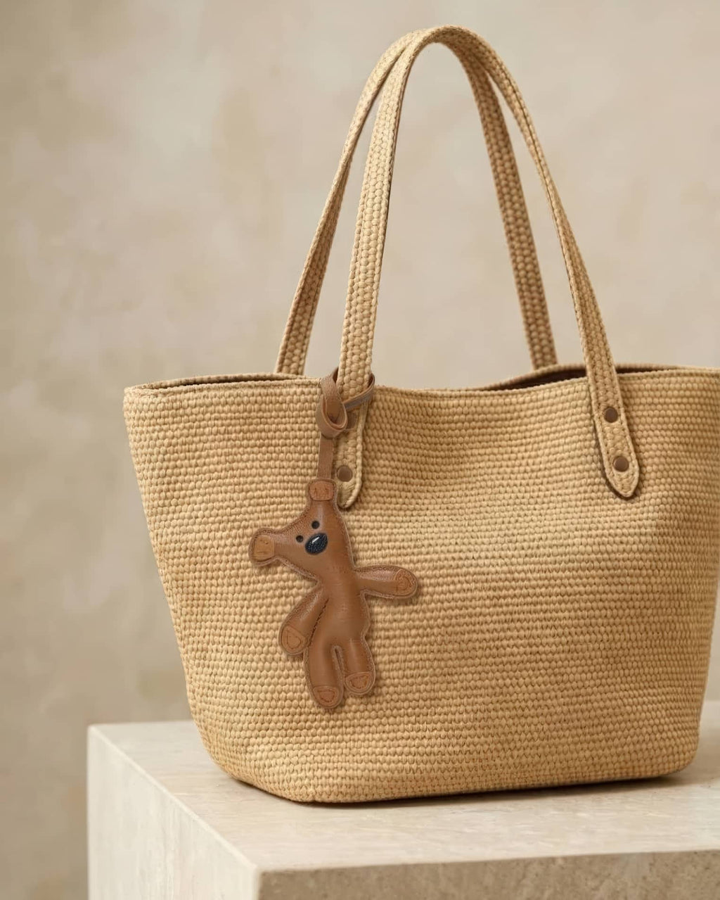 Playful brown goatskin leather teddy bear charm attached to a woven straw tote bag for handbag decoration.