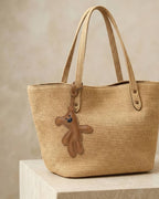 Playful brown goatskin leather teddy bear charm attached to a woven straw tote bag for handbag decoration.