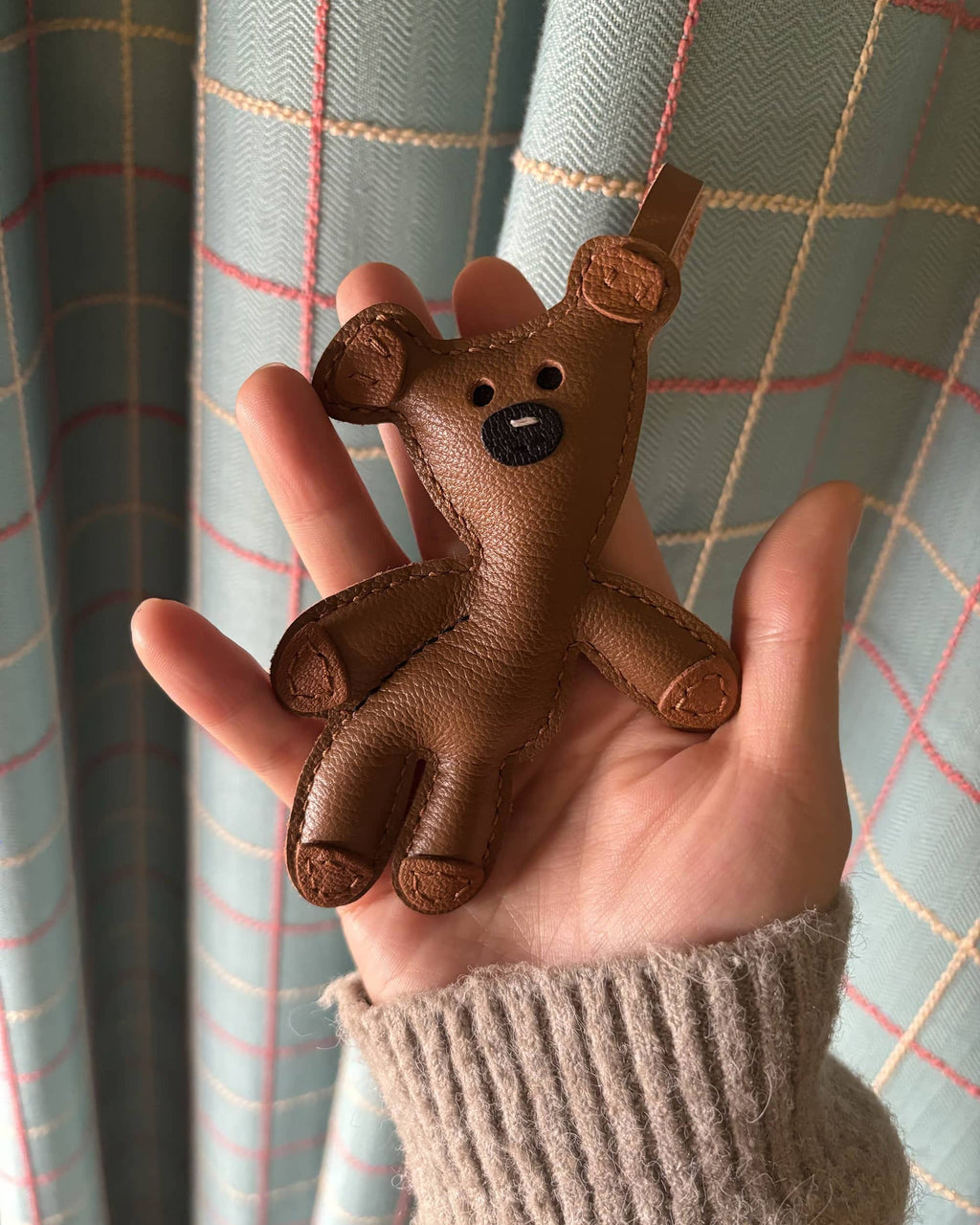 Close-up of a brown leather teddy bear bag charm held in a hand, showing fine stitching details and textured goatskin leather.
