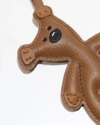Macro close-up of a brown leather teddy bear charm showing detailed embroidery facial features and edge stitching.