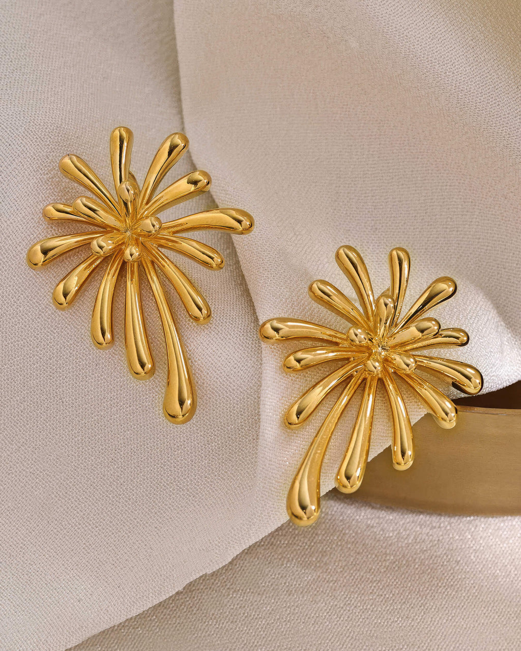 Pair of 18K gold plated burst stud earrings displayed on a soft cream fabric background, highlighting the luxury aesthetic.