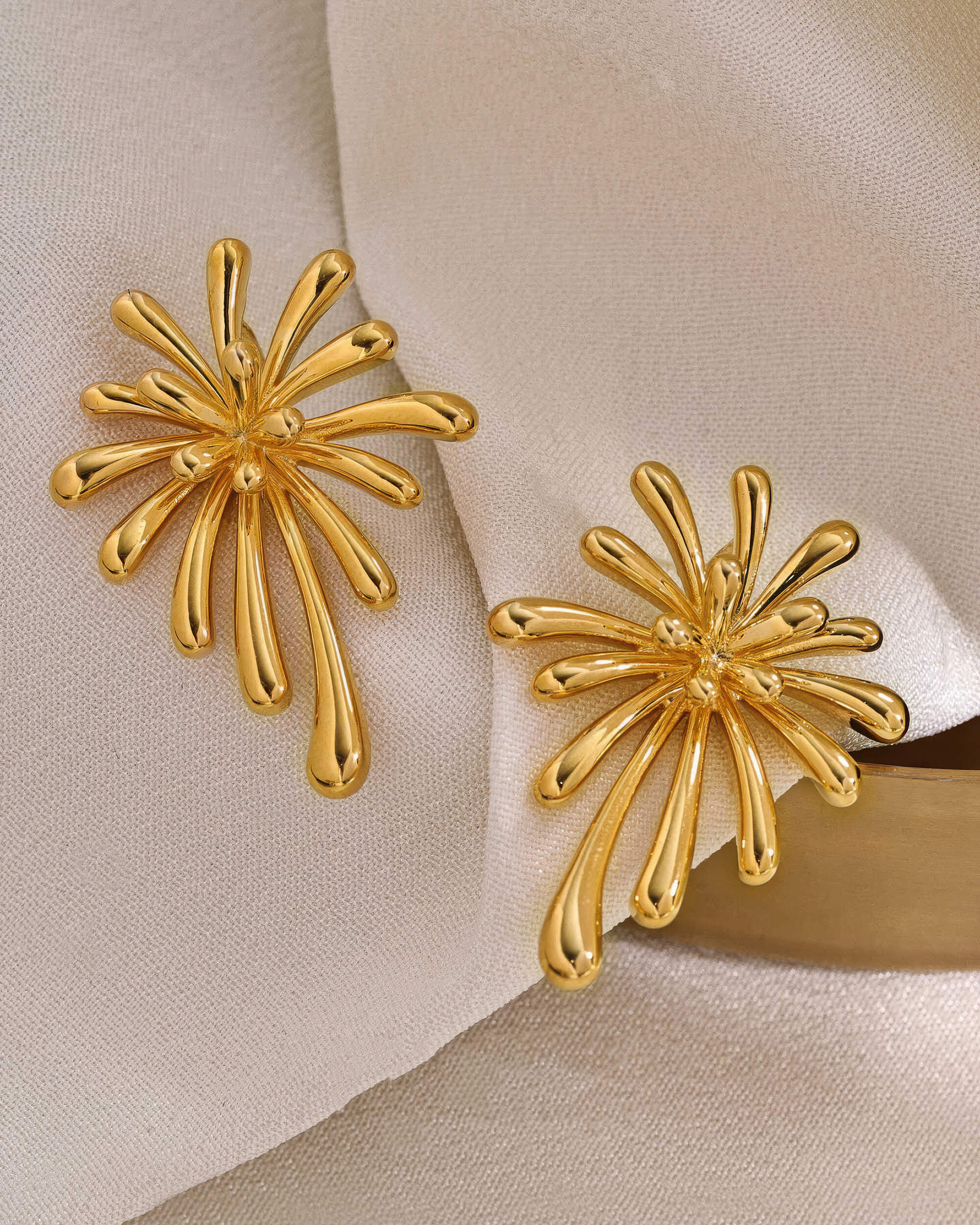 Pair of 18K gold plated burst stud earrings displayed on a soft cream fabric background, highlighting the luxury aesthetic.