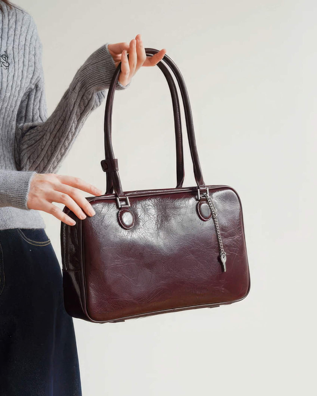 A woman holding the long straps of a burgundy distressed leather Boston bag with a silver key charm.