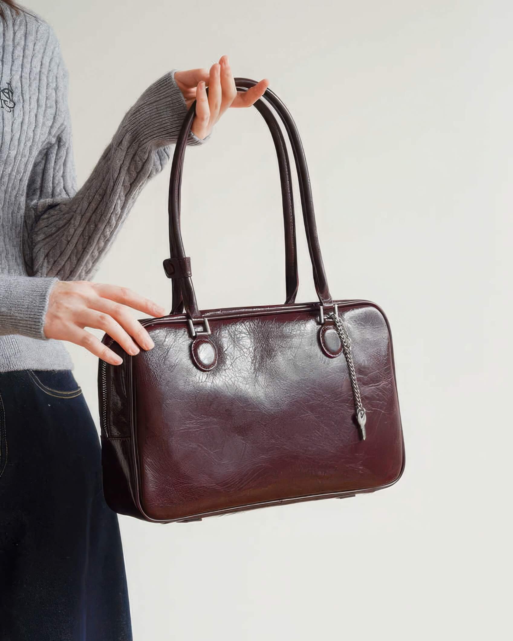 A woman holding the long straps of a burgundy distressed leather Boston bag with a silver key charm.