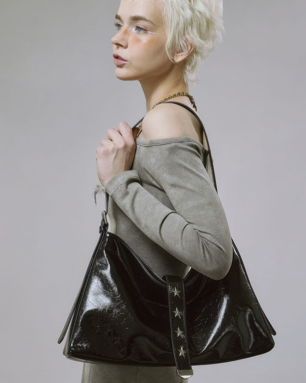 Close-up side profile of model wearing black glossy slouchy handbag featuring silver star studs on the center strap