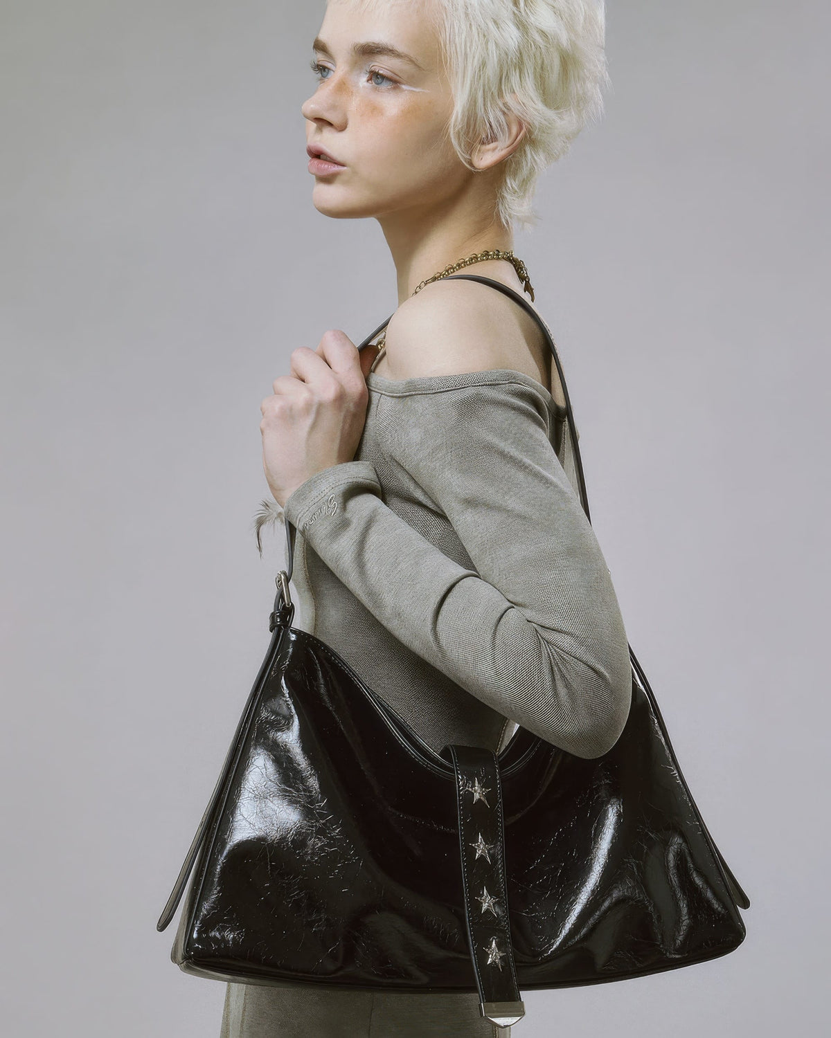 Close-up side profile of model wearing black glossy slouchy handbag featuring silver star studs on the center strap