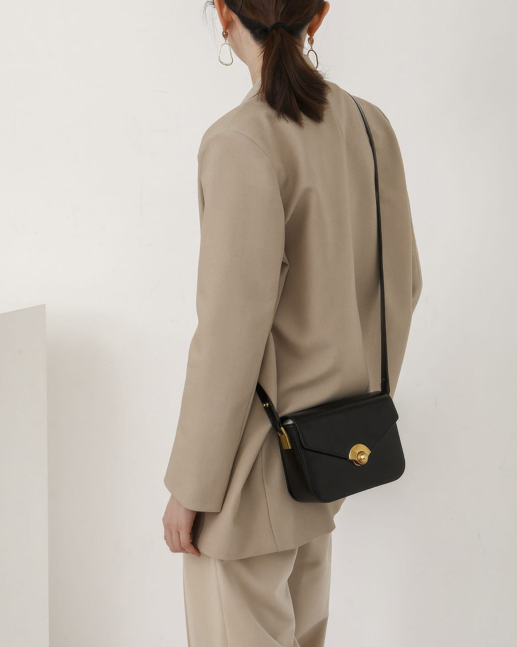Back side view of a model in a beige suit wearing a black leather flap crossbody purse.