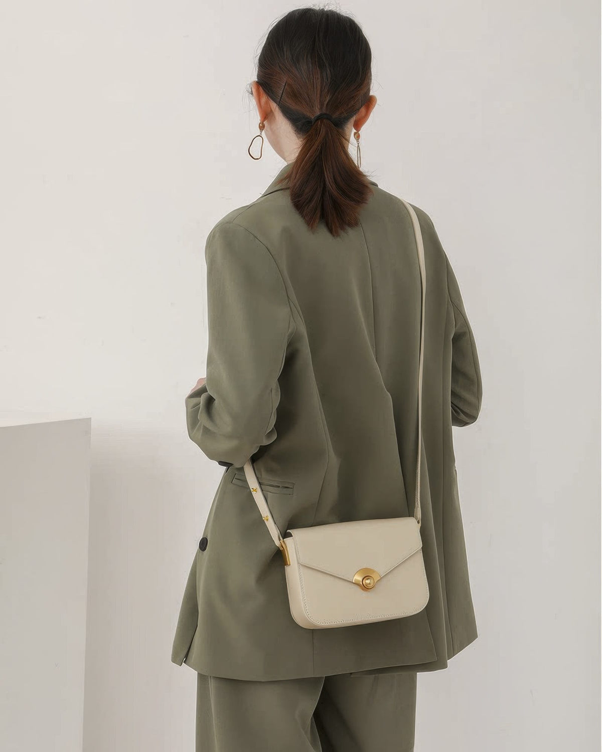 Back side view of a model in an olive green blazer outfit wearing an off-white leather flap crossbody bag.